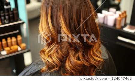 A woman sits in a salon chair, her wavy hair featuring beautiful caramel highlights. The salon is contemporary, with various hair products neatly arranged on the shelves behind her. 124047402