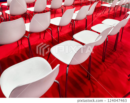 Rows of empty white chairs arranged on a vibrant red carpet for an upcoming event at a convention center Rows of empty white chairs arranged on a vibrant red carpet for an upcoming event at a convention center 124047463