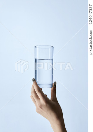 Close-up photo of hand with manicured nails holding clear glass filled with water, against light background. Mock up. 124047527