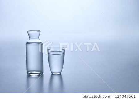 Photo of clear glass carafe and matching glass, both filled with transparent water, against soft blue background. Mock up. 124047561