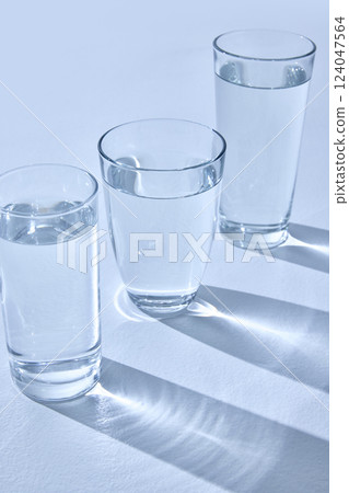 Three glasses filled with water stand on light surface, casting long, blurry shadows. Light refracts through water, creating sparkling effect. Mock up. Three glasses filled with water stand on light surface, casting long, blurry shadows. Light refracts through water, creating sparkling effect. Mock up. 124047564