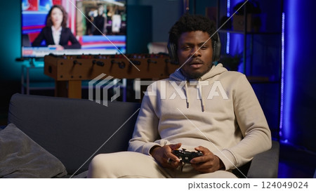African american player relaxing at home sitting down on couch, excited to play games and enjoy leisure time. Person on sofa in a decorated living room, playing video games on console. Camera A. African american player relaxing at home sitting down on couch, excited to play games and enjoy leisure time. Person on sofa in a decorated living room, playing video games on console. Camera A. 124049024