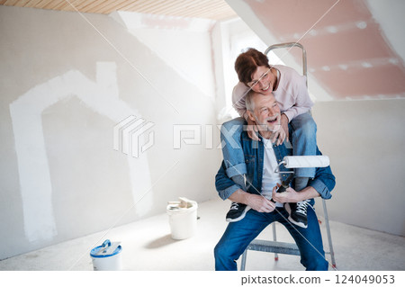 Senior husband and wife standing on a ladder repainting the walls. 124049053