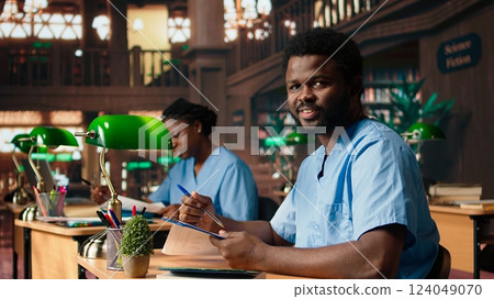 Medical assistant student reading under a green lamp in the library, doing research about medical methods and preparing for exams. Young man shows his commitment for his career. Camera A. Medical assistant student reading under a green lamp in the library, doing research about medical methods and preparing for exams. Young man shows his commitment for his career. Camera A. 124049070