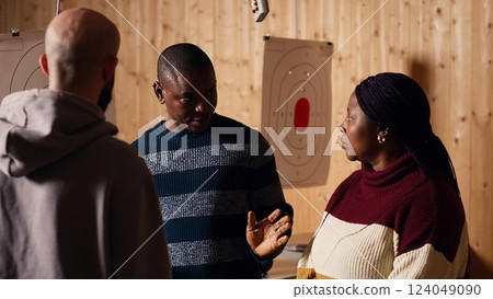 Shooting range safety officer ensuring clients are following regulations and operating firearms safely. African american firing range gallery worker giving instructions to customers, camera B 124049090