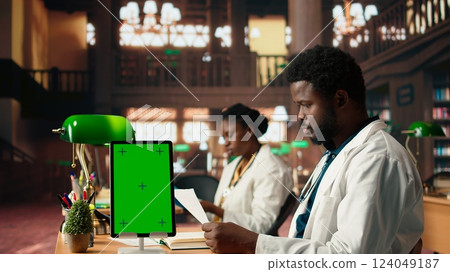 African american student reads surgery notes in a library next to mockup, studying hard for an upcoming exam. Young man emphasizes his focus on building a successful healthcare career. Camera A. 124049187