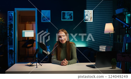 Smiling influencer entering living room used as professional studio, recording vlogs for online channel. Upbeat teenager sitting down at desk in apartment, discussing topics with audience, camera B 124049249
