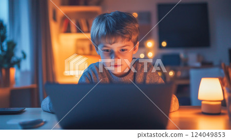 Young boy focuses intently on a laptop screen while sitting at a cozy desk in a warmly lit room during evening hours 124049853