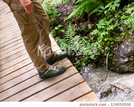 Close-up of hikers legs wearing green hiking shoes and beige cargo pants, standing at the edge of wooden bridge over muddy mountain stream. Outdoor adventure and trekking concept Close-up of hikers legs wearing green hiking shoes and beige cargo pants, standing at the edge of wooden bridge over muddy mountain stream. Outdoor adventure and trekking concept 124050500