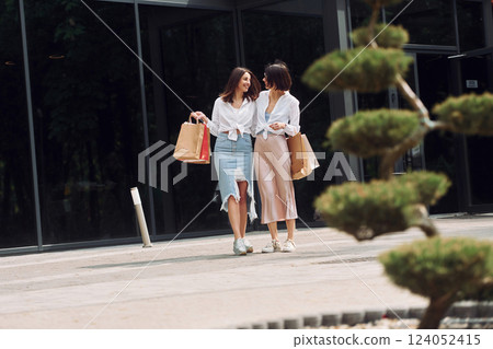 Near modern building. Two female friends have a shopping day. Walking outdoors with bags 124052415