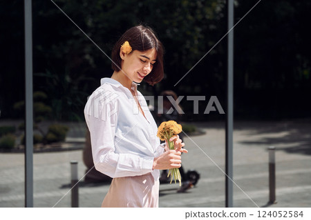 Against building with big windows. Beautiful brunette is standing outdoors with flowers in her hands Against building with big windows. Beautiful brunette is standing outdoors with flowers in her hands 124052584