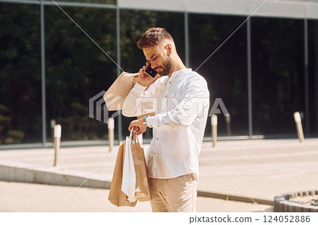 Checking the time and talking by phone. Stylish young man have shopping day. Outdoors with bags 124052886