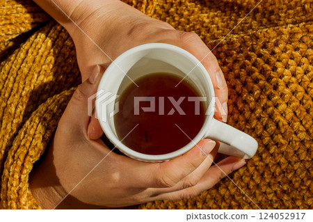 Female hands holding white tea cup in hands top view. Woman wrapped in warm brown plaid is drinking hot beverage in the morning. Cozy relax, leisure. 124052917