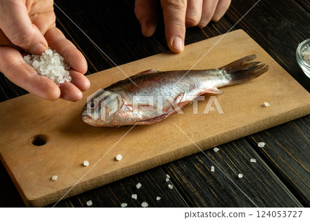 Cooking crucian carp with salt or Rutilus heckelii on a wooden kitchen board. A man's hand adds coarse salt to raw fish. 124053727