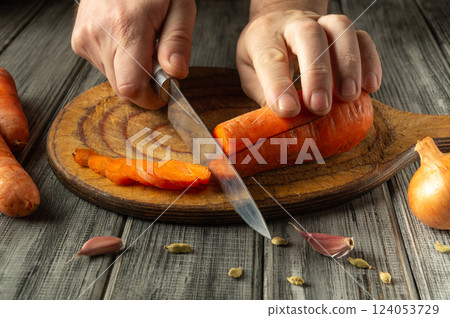 A person skillfully cuts bright orange carrots on a rustic wooden board, surrounded by garlic and cardamom pods, depicting a cozy kitchen atmosphere A person skillfully cuts bright orange carrots on a rustic wooden board, surrounded by garlic and cardamom pods, depicting a cozy kitchen atmosphere 124053729