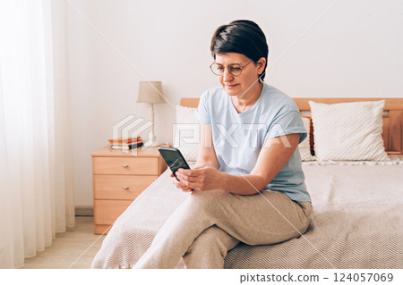 Mature caucasian woman using smartphone in cozy bedroom setting 124057069