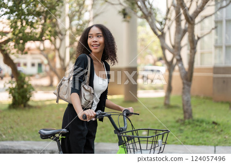 Joyful Exploration and Eco-Friendly Transport. A young woman smiles while walking with her bicycle through a sunny urban landscape. 124057496