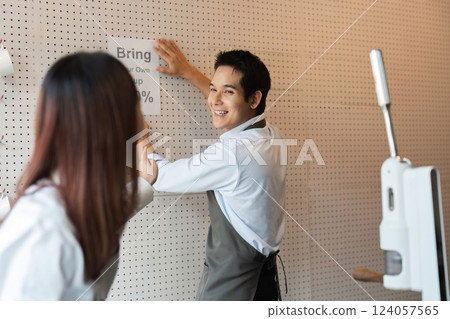 Eco-Friendly Promotion and cafe Community. A barista playfully engaging with her colleague while putting up a sign to encourage customers to bring their own cups for discounts. 124057565