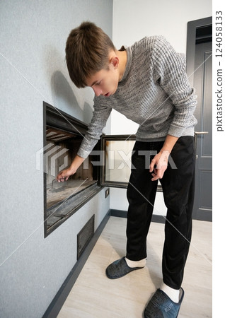 Young man tending to a modern fireplace in a contemporary living room with light gray walls and minimalistic decor while dressed in casual attire during a quiet afternoon 124058133