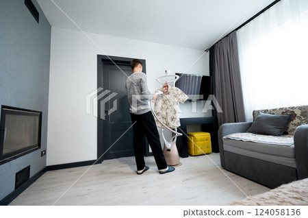 Man using a garment steamer to remove wrinkles from a floral shirt in a modern living room with elegant decor and soft natural light in the morning Man using a garment steamer to remove wrinkles from a floral shirt in a modern living room with elegant decor and soft natural light in the morning 124058136