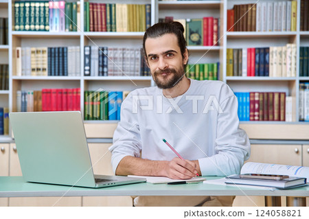 Young male teacher working in library sitting at desk with laptop books, looking at camera 124058821