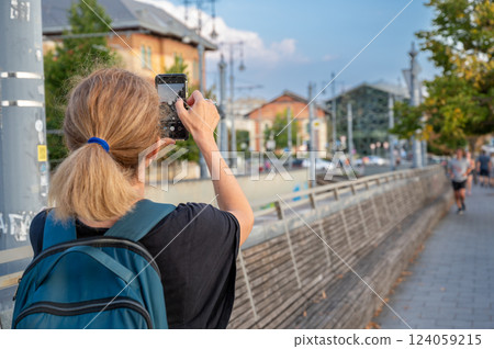 Budapest, Hungary, August 30, 2022. City life, in the background the Balna shopping center side by side with people on the Danube embankment. A middle-aged woman takes pictures with her mobile phone. 124059215