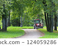 A red tractor with a trailer is parked on a gravel path in a park. Its bucket is filled with gravel. Repairing paths in a park. Preparing for the tourist season. 124059286
