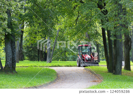 A red tractor with a trailer is parked on a gravel path in a park. Its bucket is filled with gravel. Repairing paths in a park. Preparing for the tourist season. A red tractor with a trailer is parked on a gravel path in a park. Its bucket is filled with gravel. Repairing paths in a park. Preparing for the tourist season. 124059286