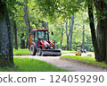 A red tractor with a trailer is parked on a gravel path in a park. Its bucket is filled with gravel. Repairing paths in a park. Preparing for the tourist season. 124059290