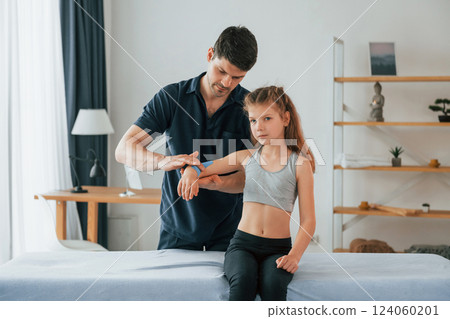 Girl is sitting. Professional physiotherapist putting kinesio tape on kid arms in clinic 124060201