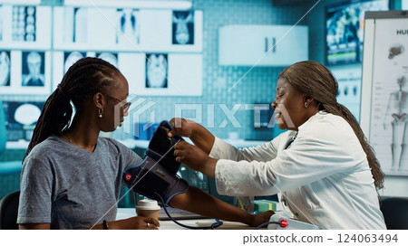 Black medic practitioner takes blood pressure with a blood pressure monitor, does a consultation with female patient using modern advanced tools for the checkup routine. Camera A. 124063494