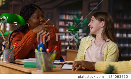 Primary school student sitting at desk in the library and reading next to mockup on tablet, preparing notes and ideas for future lessons. Scholar girl using digital gadget for assignments. Camera A. 124063510