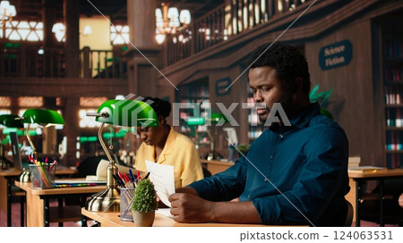 Black university student feeling anxious and struggling with learning, trying to memorize relevant information from course notes at the library. Male pupil studying to pass exams. Camera A. Black university student feeling anxious and struggling with learning, trying to memorize relevant information from course notes at the library. Male pupil studying to pass exams. Camera A. 124063531