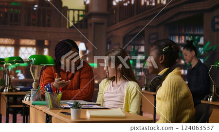 Diverse schoolgirls work with professors on homework next to mockup screen, focused on writing an essay in a school library. Scholars completing their assignment on textbooks. Camera A. Diverse schoolgirls work with professors on homework next to mockup screen, focused on writing an essay in a school library. Scholars completing their assignment on textbooks. Camera A. 124063537