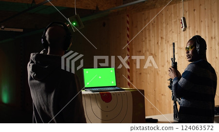 Green screen laptop next to shooting gallery safety officer watching client shoot at targets, supervising him. Mockup notebook and firing range expert teaching man how to use firearms, camera A 124063572