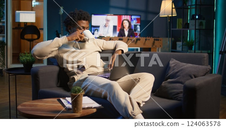 Positive african american man multitasking on a laptop in stylish loft, fully connected to the internet while managing his freelance work and remote responsibilities. Browsing web at home. Camera B. 124063578