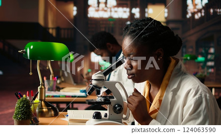 Female science student examines cells under the microscope for her scientific research in a campus library, takes notes about chemical processes and reactions for her thesis. Camera A. Female science student examines cells under the microscope for her scientific research in a campus library, takes notes about chemical processes and reactions for her thesis. Camera A. 124063590