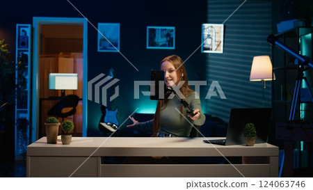 Girl in neon lit apartment using phone on selfie stick to capture welcoming intro for vlogging channel. Joyful entertainer in living room connecting with loyal audience during live stream, camera B 124063746