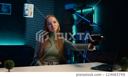Upbeat teenager films content for online streaming channel in apartment studio using phone on selfie stick. Smiling gen z influencer at home interacts with subscribers, recording with phone, camera B Upbeat teenager films content for online streaming channel in apartment studio using phone on selfie stick. Smiling gen z influencer at home interacts with subscribers, recording with phone, camera B 124063749