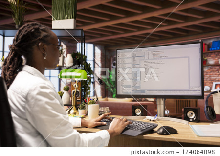 Woman in home office looking at UI on computer screen, typing and executing programming code. Coding application UI used by african american IT developer writing and testing code in modern apartment 124064098