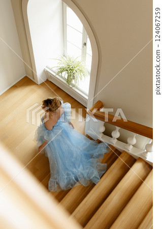 Young woman in light blue evening dress climbs large wooden staircase in luxurious manor. Top view. 124067259