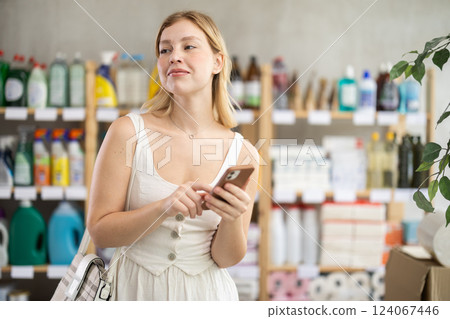 Female shopper searches for necessary goods on the shelves of a supermarket, checking list on her smartphone Female shopper searches for necessary goods on the shelves of a supermarket, checking list on her smartphone 124067446