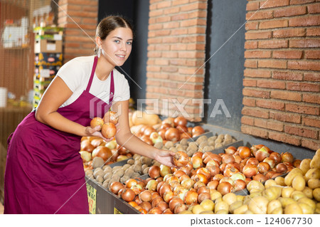 Girl seller of garden stuff store is replenishing showcase with vegetables, puts onion on showcase 124067730