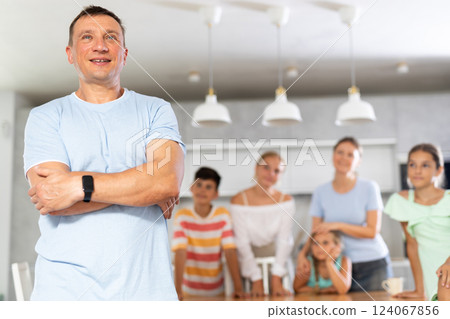 Smiling dad poses in cozy kitchen, large family with four children and wife are standing behind Smiling dad poses in cozy kitchen, large family with four children and wife are standing behind 124067856