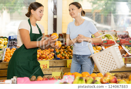 Polite salesgirl offering sweet oranges to female customer in greengrocery store 124067883