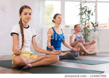 Middle-aged woman, man, preteen girl and boy practicing Padmasana with crossed legs on mats during family Hatha yoga class in wellness studio 124067990