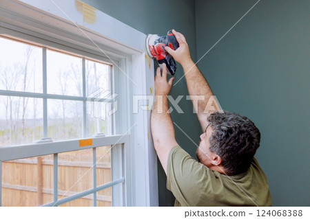 Skilled worker is sanding wooden window trim, preparing for painting in home improvement effort. 124068388