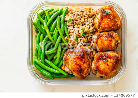 Flat lay of a meal prep container featuring baked chicken thighs, brown rice, and fresh green beans, arranged in a glass container on a white background 124069577