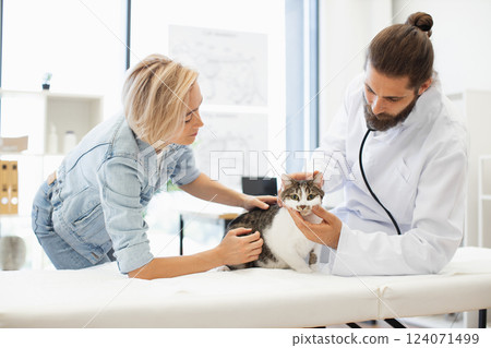 Male veterinarian using stethoscope to check health of tabby cat, female owner assisting, in modern clinic. Image showcasing healthcare, animals, compassion, and care. Male veterinarian using stethoscope to check health of tabby cat, female owner assisting, in modern clinic. Image showcasing healthcare, animals, compassion, and care. 124071499