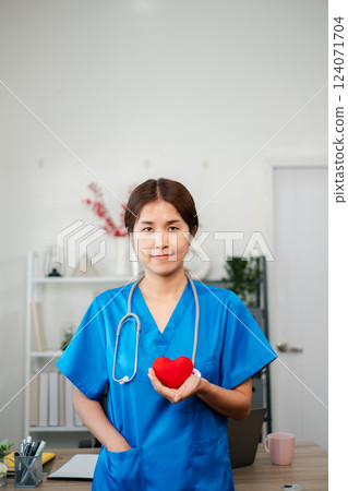 A nurse in blue scrubs holds a red heart symbol, standing in a modern medical office environment. 124071704